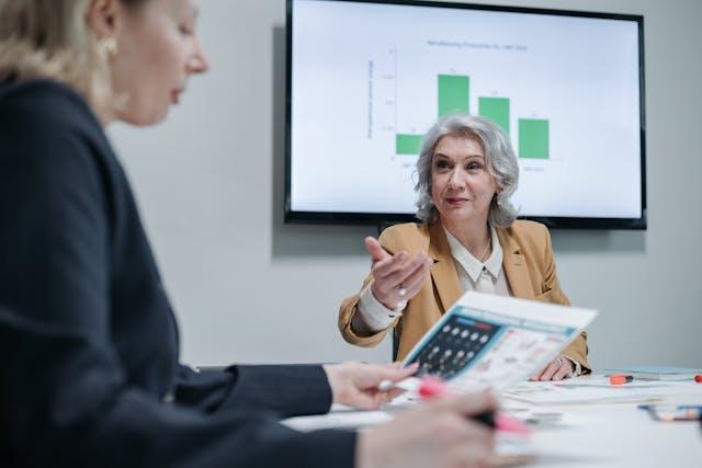A woman in a beige suit explains compliance matrix details to a woman in a blue suit while a presentation runs in the background