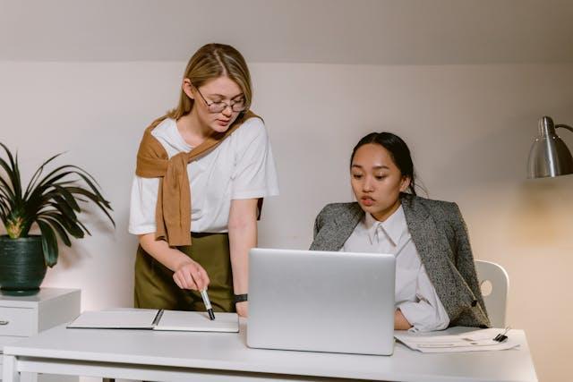 Woman in white shirt standing and helping woman in gray blazer prepare a compliance matrix - BIT Solutions LLC Woman in white shirt standing and helping woman in gray blazer prepare a compliance matrix