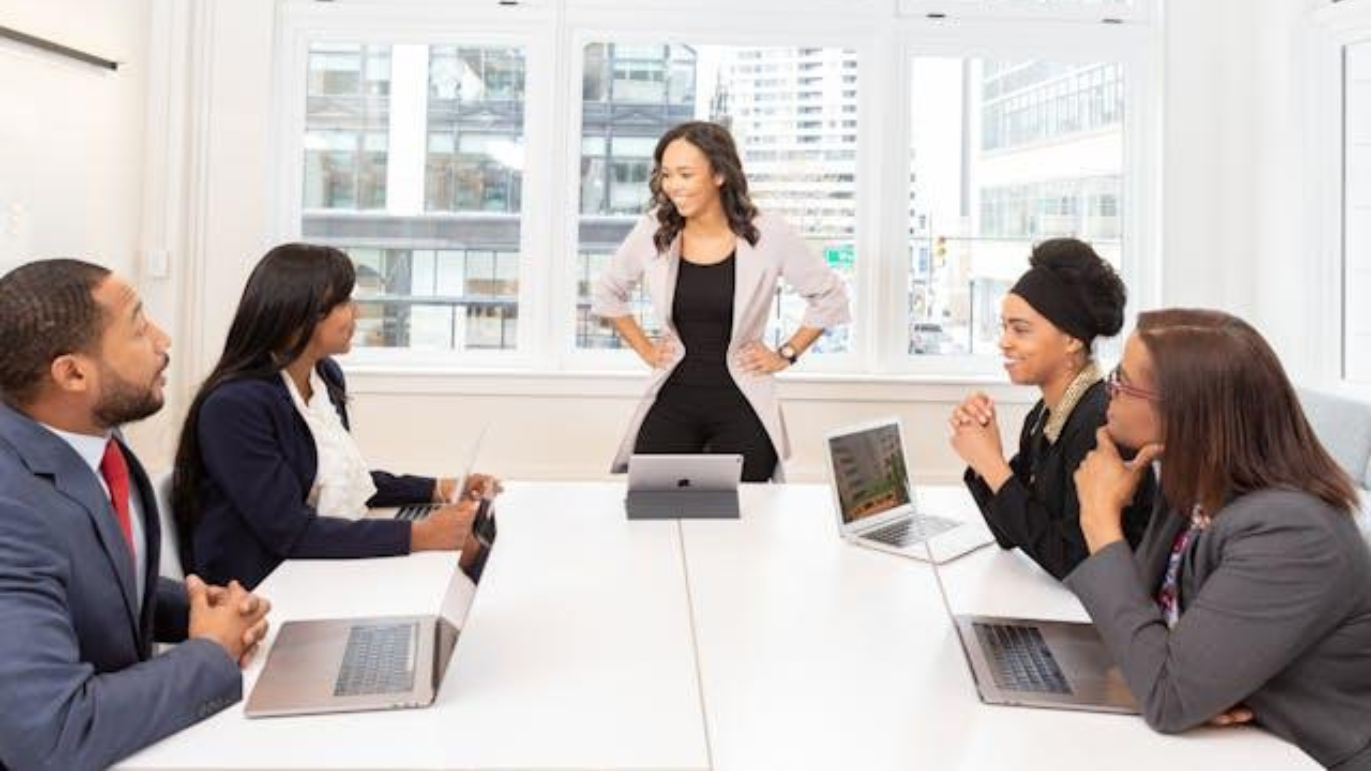 A confident woman leads a business meeting in a bright modern office as four colleagues listen attentively around a white conference table