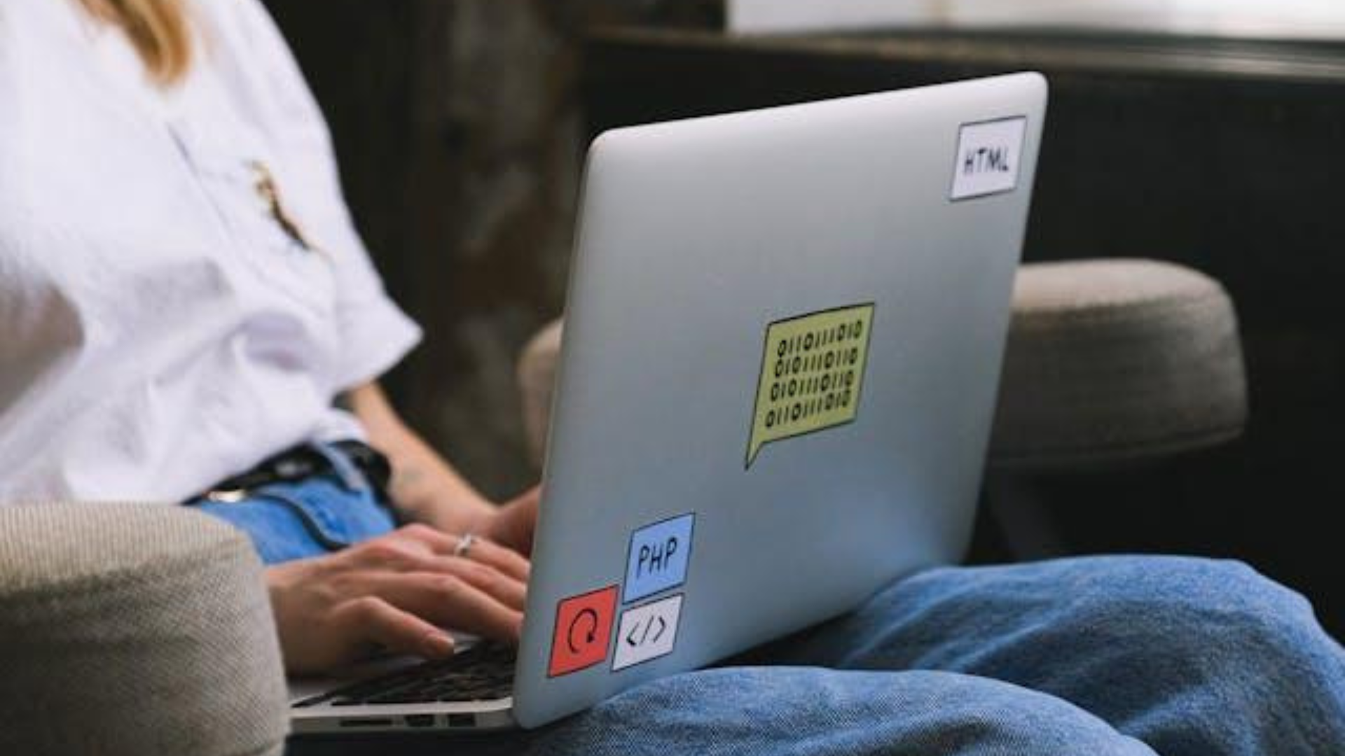 A person typing on a silver laptop featuring coding-themed stickers