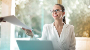 A smiling professional woman in a white blazer and glasses receives a document during a meeting