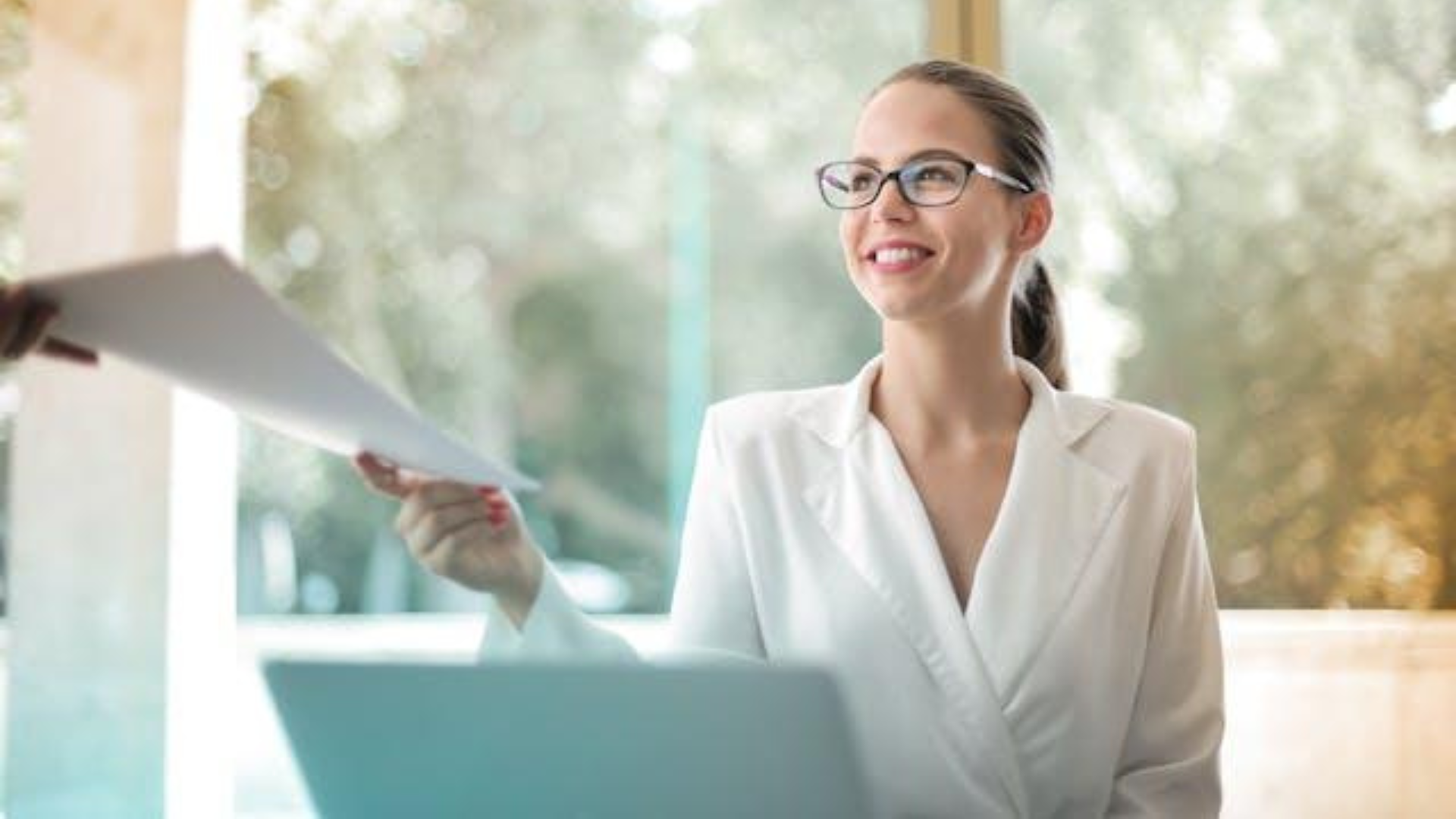 A smiling professional woman in a white blazer and glasses receives a document during a meeting