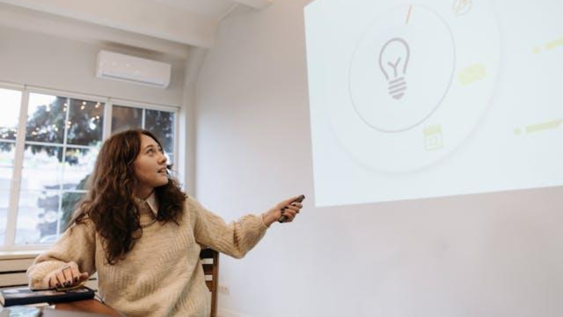 A woman with long curly hair points a remote at a large wall projection of a lightbulb icon and data graphics during a creative presentation in a brightly lit room